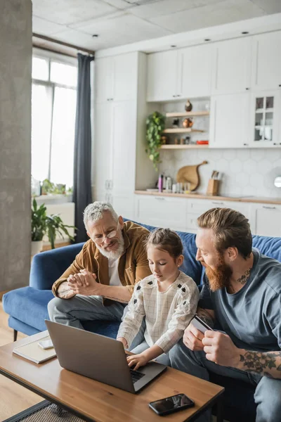 Gay father holding credit card while daughter and partner using laptop at home — Stock Photo