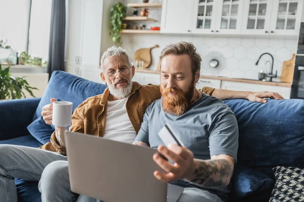 Tattooed gay man holding credit card and using laptop near partner with coffee at home — Stock Photo