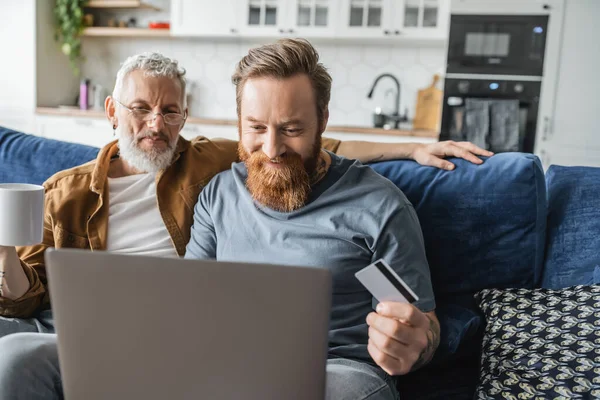Smiling homosexual man holding credit card near laptop and partner with coffee at home — Stock Photo