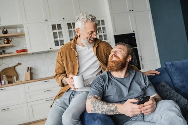 Smiling gay man holding coffee cup near partner with smartphone on couch at home — Stock Photo