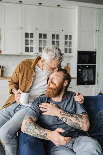 Reifen gay Mann halten Kaffee und küssen fröhlich Partner mit smartphone in Wohnzimmer — Stockfoto
