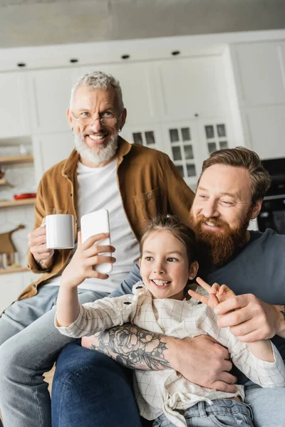 Smiling girl showing peace gesture while taking selfie with homosexual fathers at home — Stock Photo