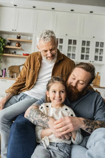 Homosexuelle Eltern umarmen sich und schauen lächelnde Tochter zu Hause an — Stockfoto