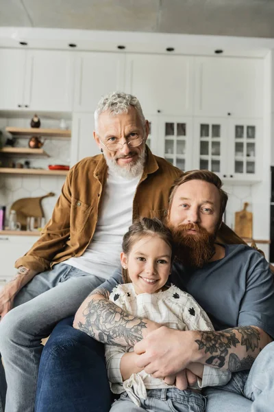 Homosexueller Mann umarmt lächelnde Tochter in der Nähe seines Partners und schaut zu Hause in die Kamera — Stockfoto