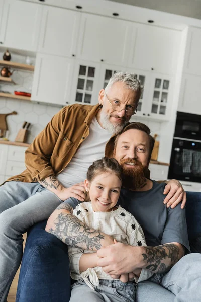 Los padres del mismo sexo sonriendo y abrazando a la hija adoptiva en casa - foto de stock