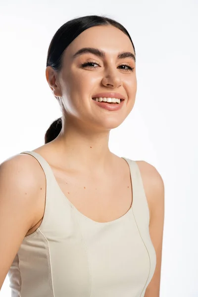 Mujer joven feliz con el pelo morena, belleza natural y piel perfecta mirando a la cámara mientras posando en camiseta y sonriendo aislado sobre fondo blanco - foto de stock