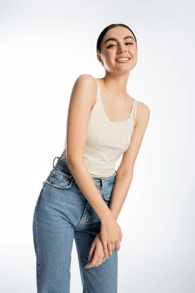 Lovely and smiling woman with brunette hair, natural beauty and perfect skin looking at camera while posing in tank top and denim jeans isolated on white background — Stock Photo