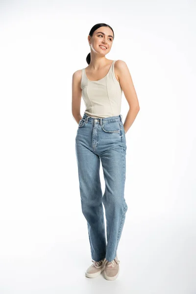 Full length of gorgeous young woman with natural makeup, brunette hair and perfect skin smiling while posing in denim jeans with tank top on white background — Stock Photo