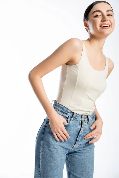Appealing and young woman with natural makeup, brunette hair and perfect skin smiling while posing in tank top and denim jeans on white background — Stock Photo