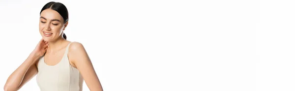 Schüchterne junge Frau mit natürlichem Make-up, brünetten Haaren und perfekter Haut, lächelnd im Tank-Top stehend, wegschauend und mit der Hand am Hals isoliert auf weißem Hintergrund posierend, Banner — Stockfoto