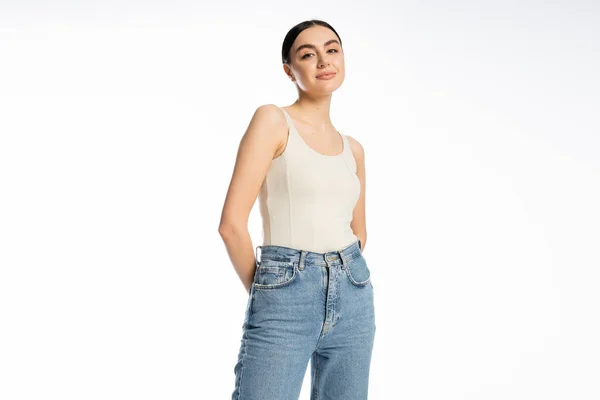 Appealing and young woman with natural makeup, brunette hair and perfect skin smiling while posing in tank top and denim jeans and looking at camera on white background — Stock Photo