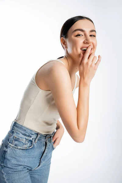 Shy woman with natural beauty, brunette hair and perfect skin laughing and covering mouth while posing in tank top and denim jeans and looking at camera on white background — Stock Photo