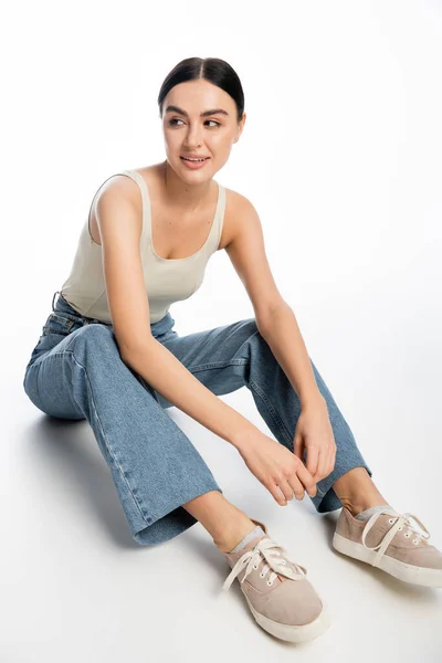 Full length of pleased young woman with natural makeup, brunette hair and perfect skin smiling while sitting in denim jeans and tank top while looking away on white background — Stock Photo