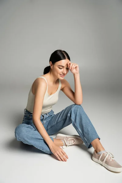 Full length of charming young woman with natural makeup, brunette hair and perfect skin smiling while sitting in denim jeans and tank top while looking down on grey background — Stock Photo