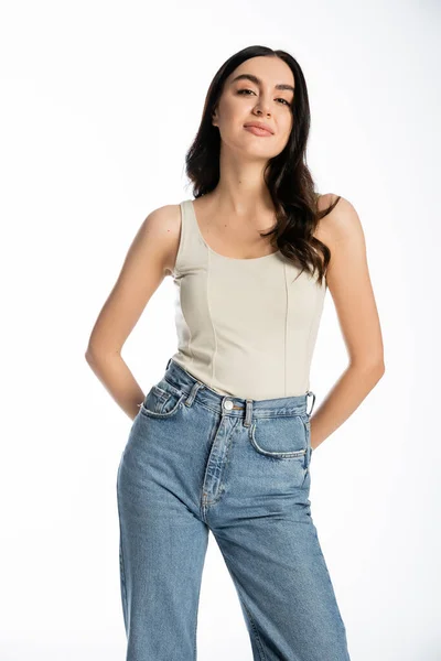 Charming woman with natural beauty, brunette hair and perfect skin smiling while posing in denim jeans and tank top and looking at camera on white background — Stock Photo