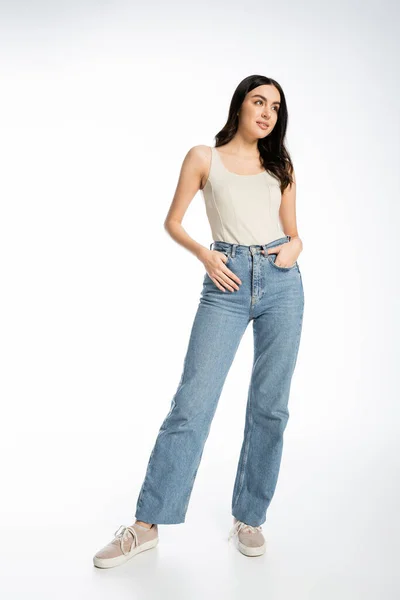 Full length of enchanting woman with natural beauty, brunette hair and perfect skin smiling while posing with hand in pocket while standing in denim jeans and tank top on white background — Stock Photo