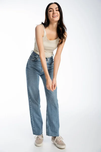 Full length of enchanting woman with brunette hair, perfect skin and natural makeup standing in denim jeans and tank top while smiling and looking at camera on white background — Stock Photo