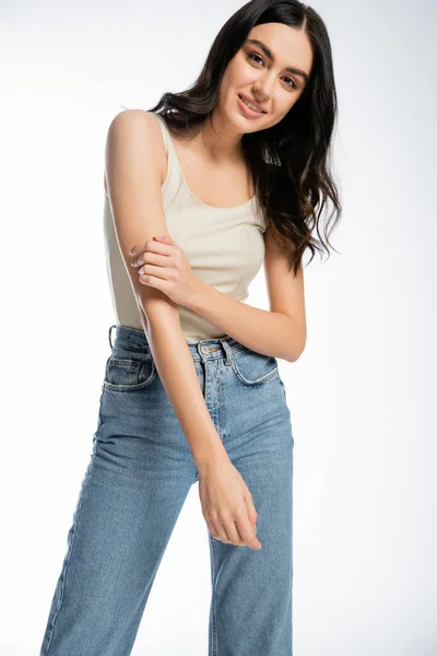 Enchanting woman with brunette hair, perfect skin and natural makeup standing in denim jeans and tank top while smiling and looking at camera on white background — Stock Photo
