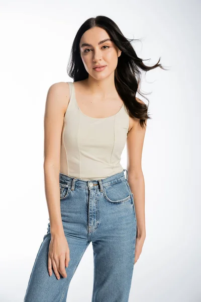Gorgeous woman with brunette hair, perfect skin and natural makeup standing in denim jeans and tank top while looking at camera on white background — Stock Photo