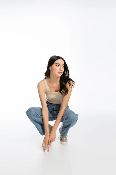 Volle Länge der jungen und attraktiven Frau mit natürlichem Make-up, brünetten Haaren und perfekter Haut sitzt in Jeans und Tank-Top und schaut weg auf weißem Hintergrund — Stockfoto