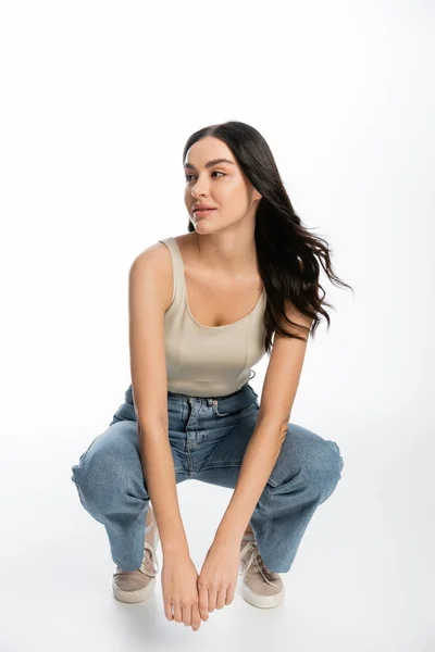 Full length of young and carefree woman with natural makeup, brunette hair and perfect skin sitting in denim jeans and tank top and looking away on white background — Stock Photo