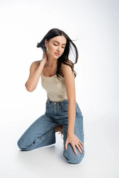 Full length of young and alluring woman with natural makeup, brunette hair and perfect skin standing on knees and posing in denim jeans with tank top while looking away on white background — Stock Photo