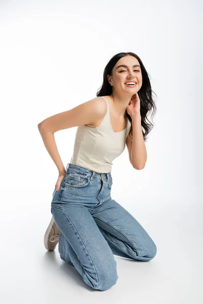 Full length of young and joyful woman with natural makeup, brunette hair and perfect skin standing on knees and posing in denim jeans with tank top while looking away on white background — Stock Photo
