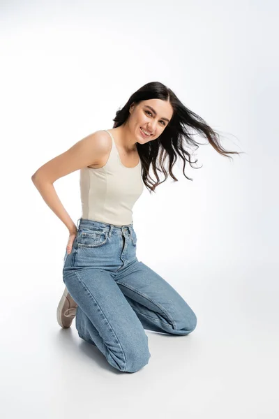 Full length of young and gorgeous woman with flawless makeup, brunette hair and perfect skin standing on knees and posing in denim jeans with tank top while on white background — Stock Photo