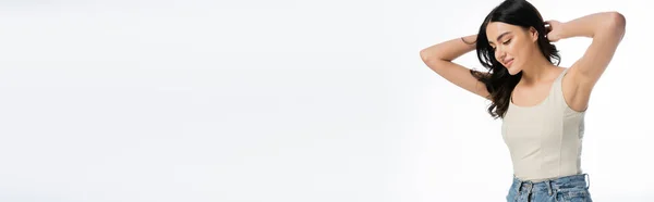 Mujer joven y seductora con maquillaje natural, cabello moreno y piel perfecta posando en camiseta con las manos detrás de la cabeza y sonriendo aislada sobre fondo blanco, pancarta - foto de stock