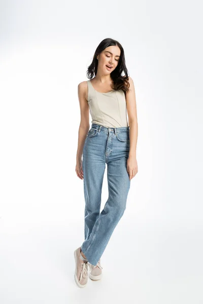 Full length of alluring woman with opened mouth, natural makeup, brunette hair and perfect skin posing in tank top and denim jeans on white background — Stock Photo