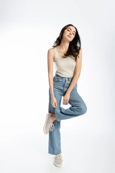 Full length of alluring woman with shiny brunette hair, natural makeup and perfect skin posing in tank top and touching denim jeans on white background — Stock Photo