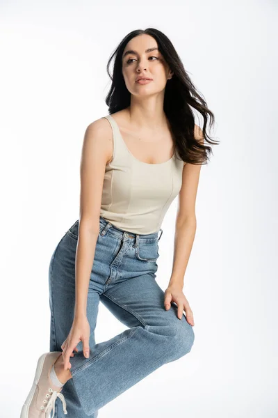 Чудова та молода жінка з блискучим волоссям брюнетки, натуральним макіяжем та ідеальною шкірою, що позує у верхній частині бака та торкається джинсових джинсів, дивлячись на білий фон — стокове фото
