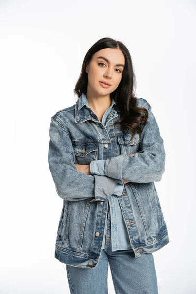 Alluring young woman with gorgeous brunette hair posing with folded arms while standing in denim jacket and looking at camera on white background — Stock Photo