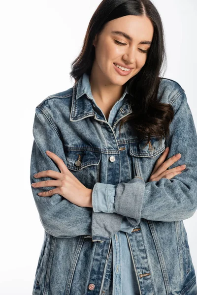 Счастливая и молодая женщина с великолепными волосами брюнетки позировать со сложенными руками, стоя в синей джинсовой куртке и глядя вниз на белом фоне — стоковое фото
