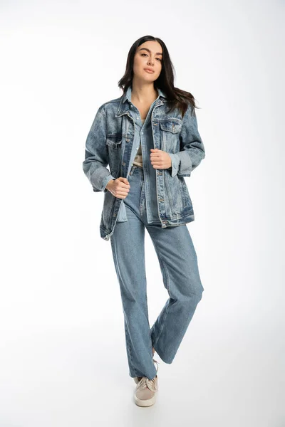Повна довжина привабливої молодої моделі з брюнеткою волосся, бездоганний і природний макіяж позує в джинсовій куртці і блакитних джинсах, стоячи і дивлячись на камеру на білому тлі — стокове фото