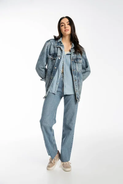 Полная длина модной молодой модели с брюнеткой волосы и безупречный макияж позируя в джинсовой куртке и синих джинсах, стоя и глядя на камеру на белом фоне — стоковое фото