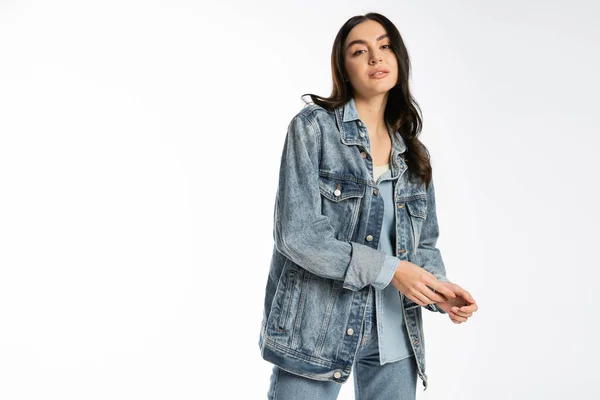 Модная молодая модель с брюнетками и безупречным макияжем позируя в синей джинсовой куртке и джинсах стоя и глядя на камеру на белом фоне — стоковое фото