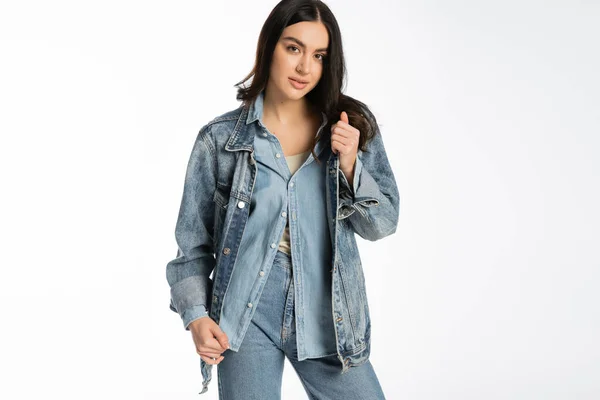 Стильная молодая модель с брюнетками и безупречным макияжем, позирующая в модной джинсовой куртке и синих джинсах, стоя и глядя на камеру на белом фоне — стоковое фото