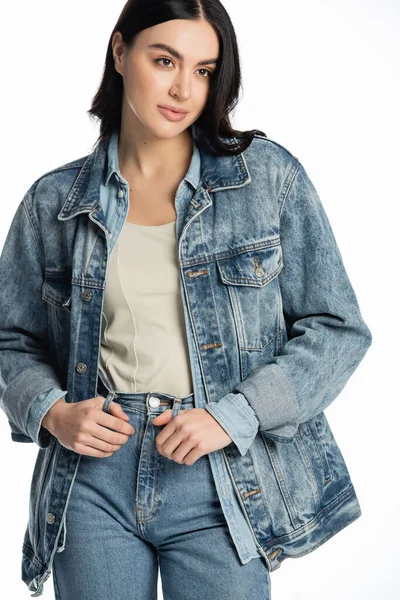 Young and fashionable model with brunette hair and natural makeup posing in stylish denim outfit while standing and looking away on white background — Stock Photo