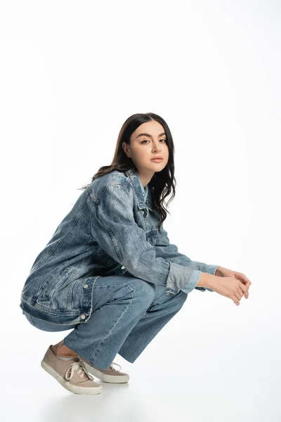 Full length of gorgeous young model with brunette hair and natural makeup posing in trendy denim outfit while sitting on haunches and looking at camera on white background — Stock Photo