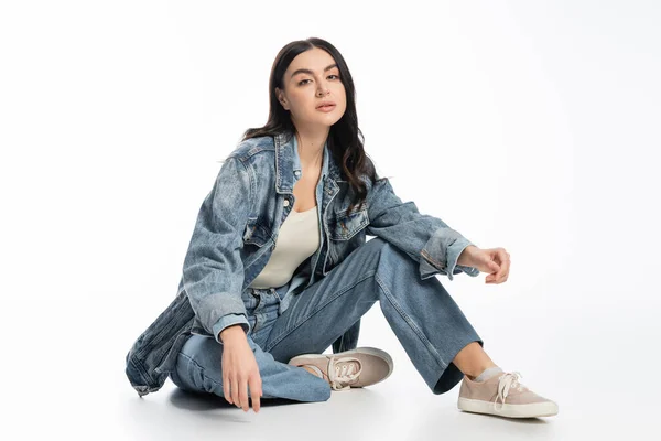 Full length of chic young model with brunette hair and natural makeup posing in trendy denim outfit while sitting and looking at camera on white background — Stock Photo