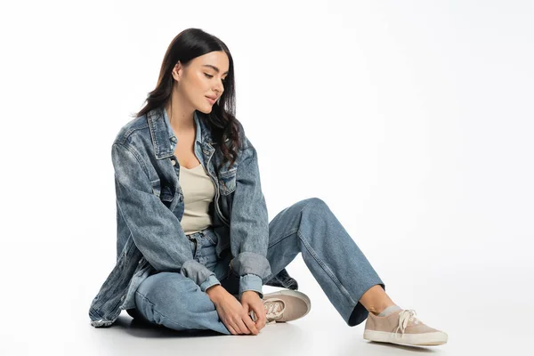 Full length of alluring young woman with natural makeup and brunette hair posing in trendy denim outfit while sitting and looking away on white background — Stock Photo