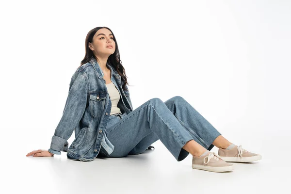Full length of dreamy young woman with natural makeup and brunette hair posing in trendy denim outfit while sitting and thinking, looking away on white background — Stock Photo