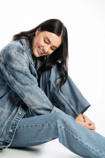 Полная длина заманчивой женщины с натуральным макияжем и брюнеткой здоровые волосы позируют в модном джинсовом наряде, сидя и улыбаясь на белом фоне — стоковое фото