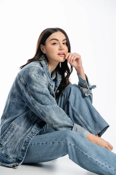 Attractive young woman with gorgeous brunette hair posing in stylish denim jacket and jeans while sitting and holding finger near lips on white background — Stock Photo