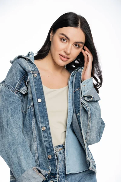Enchanting young woman with gorgeous brunette hair posing in stylish blue denim jacket and holding hand near face while looking at camera on white background — Stock Photo