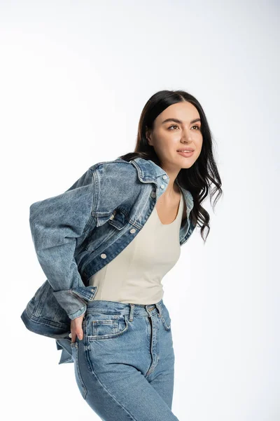 Dreamy young woman with gorgeous brunette hair posing with hand in pocket of blue jeans while standing in denim jacket and looking away on white background — Stock Photo