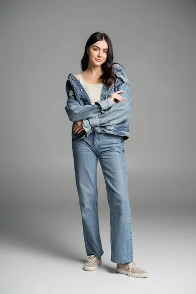Full length of alluring woman with brunette hair posing with folded arms, standing in stylish blue jeans and denim jacket and smiling while looking at camera on grey background — Stock Photo