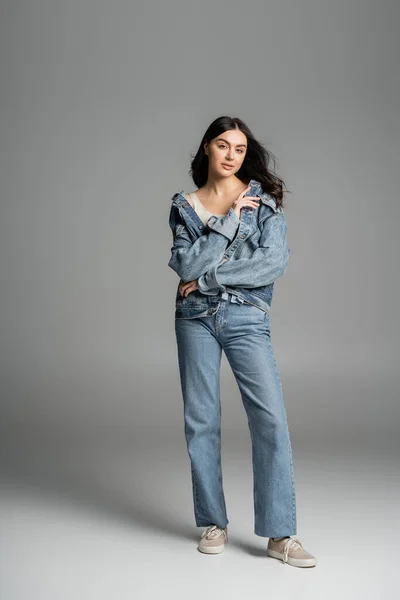 Full length of charming woman with brunette hair posing while standing in stylish blue jeans and trendy denim jacket and looking at camera on grey background — Stock Photo
