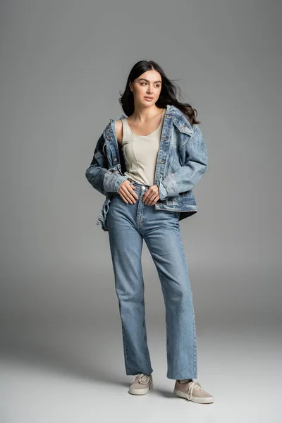 In voller Länge bezauberndes junges Model mit brünetten Haaren posiert in stylischer blauer Jeans und Jeansjacke und schaut auf grauem Hintergrund weg — Stockfoto